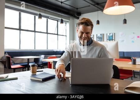 Mann lächelt, während er einen silbernen Laptop am offenen Bürotisch benutzt, der Kaffeetasse und Stift hält Stockfoto
