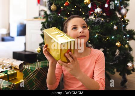Weißes Mädchen, das auf dem Boden im Wohnzimmer am Weihnachtsbaum sitzt und ein goldgewickeltes Geschenk ins Ohr hält Stockfoto