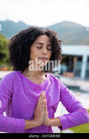 Frau im mittleren Alter, die Yoga auf der Terrasse am Pool vor dem modernen Haus übt, mit Blick auf die Berge Stockfoto