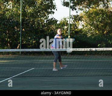 Junge Erwachsene Frau, die Schläger schwingt und Tennisball über dem Netz auf dem Outdoor Court im Park spielt Stockfoto