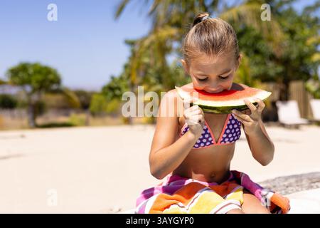 Kind Mädchen beißt große Wassermelonenscheibe, während es auf bunten Handtuch auf Bank im Garten sitzt Stockfoto