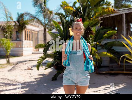 African American woman standing on tropical sandy beach wearing denim shorts holding striped towel Stockfoto