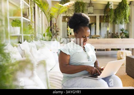 Reife afroamerikanische Frau, die im Sonnenraum auf einem Laptop schreibt, mit Couch, hängenden Pflanzen, Kopierraum Stockfoto