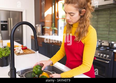 Junge Erwachsene Frau, die Paprika im Sieb unter schwarzem Wasserhahn auf der Kücheninsel wäscht, Kopierraum Stockfoto