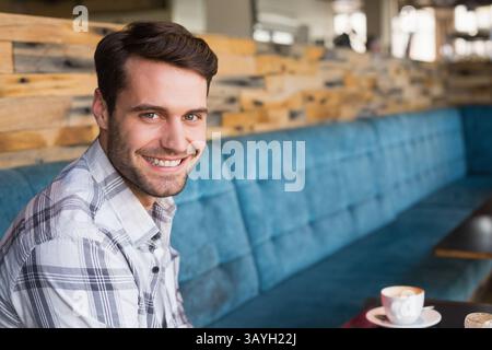 Mann sitzt auf blauer Bank im Café und lächelt in die Kamera und hält Keramikkaffee, Kopierraum Stockfoto