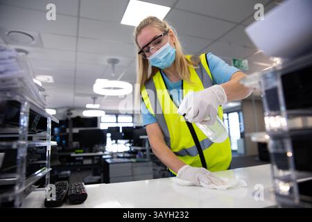 Frau trägt Sicherheitsweste Reinigungsbüroschreibtisch mit Desinfektionsflasche und Tuch im modernen Büro Stockfoto