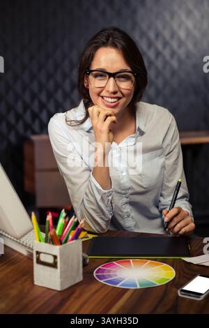 Frau, die am Schreibtisch in einem modernen Studio sitzt und den Stift über einem Grafiktablett mit Farbrad hält Stockfoto