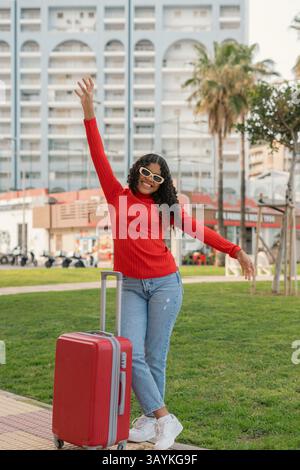 Junge schwarze Frau mit lockigen Haaren und Sonnenbrille, die glücklich winkte, mit einem Arm angehoben, während sie sich mit dem anderen auf ihrem roten Wagen in einem Stadtpark mit seinem Arm lehnte Stockfoto
