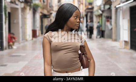 Modische junge Frau mit langen schwarzen Haaren, die ein mokkafarbenes Oberteil und eine weiße Hose trägt, die mit einer braunen Geldbörse eine Stadtstraße hinunterläuft Stockfoto