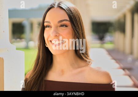 Schöne hispanische Frau mit langen braunen Haaren und Mokka-Lippenstift lächelnd und posierte an einem sonnigen Tag im Freien mit einem Mokkafarben von der Schulter Stockfoto