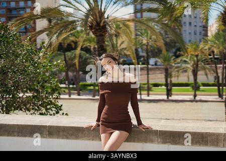Glückliche junge Frau, die ein mokkafarbenes Minikleid aus der Schulter trägt, lächelt in einem Park mit Palmen an einem sonnigen Tag und genießt Sommerferien in A Stockfoto