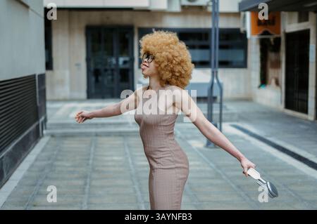 Eine junge Frau mit großen lockigen blonden Afro-Haaren, in einem beigen Kleid und einer Brille, hebt ihre Arme und hält ihre silbernen Schuhe Stockfoto