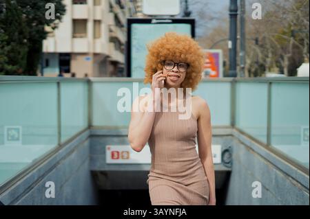 Junge schwarze Frau mit gefärbtem Afro-Haar und stilvoller Brille lächelt und plaudert auf ihrem Smartphone, während sie eine geschäftige U-Bahn-Station verlässt Stockfoto