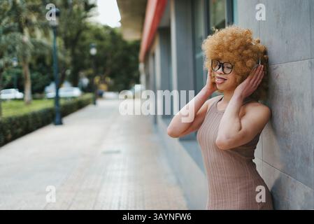 Stilvolle junge Frau mit lockigem blondem Haar und Brille genießt Musik mit Kopfhörern Stockfoto