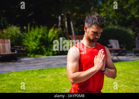 Sportlicher Mann kniet auf Gras im Garten neben Holzdeck, drückt Hände in Gebetsposition Stockfoto