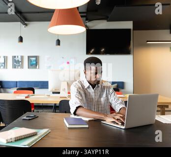 Junger afroamerikanischer Mann, der auf silbernem Laptop am Coworking Desk tippt, mit Notebooks und Smartphone Stockfoto