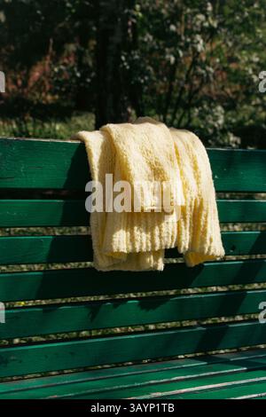 Gelbe Strickjacke aus Baumwolle auf grüner Bank. Weicher gelber Strickpullover auf der Parkbank. Großstadtmode. Strickmode für den Sommer. Gemütliches Hobby. Stockfoto