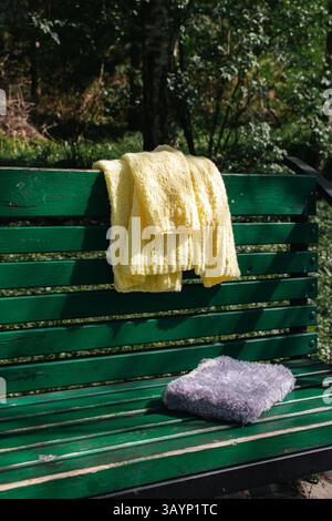 Gelbe Strickjacke aus Baumwolle und grauer Bucheinband auf grüner Bank. Weicher gelber Strickpullover und flauschiges gestricktes Buch auf Parkbank. Großstadtmode Stockfoto