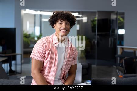 Junger Mann, der auf einem grauen Sofa in der modernen Bürolounge sitzt, mit Glastrennwand und LED-Lampen Stockfoto