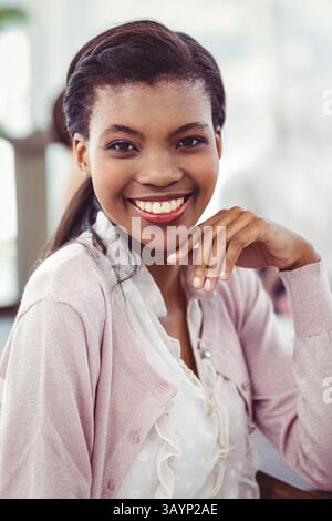 Junge Erwachsene afroamerikanische Frau, die im Büro sitzt, in weißer Bluse und rosa Strickjacke lächelnd Stockfoto