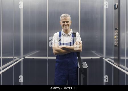 Professioneller Techniker, der mit überkreuzten Armen im Aufzug steht und die Kamera anlächelt Stockfoto