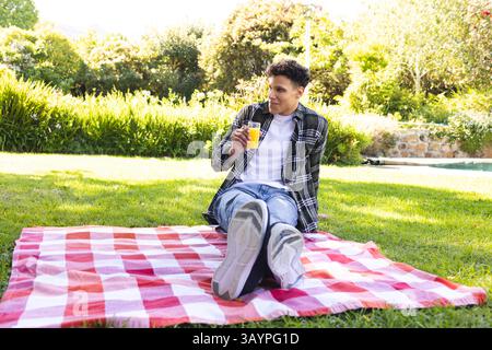 Mann sitzt auf rot-weiß karierter Picknickdecke im Garten und hält ein Glas Orangensaft Stockfoto