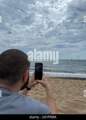 Der Tourist macht Fotos vom Meer auf seinem Telefon, das am Ufer sitzt, vertikales Foto. Hochwertige Fotos Stockfoto