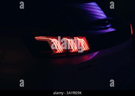 Rücklicht eines modernen Autos bei Nacht. Nahaufnahme des links leuchtenden LED-Rücklichts mit drei Streifen, lila Neonlinien, die sich in der Karosserie widerspiegeln. Stockfoto