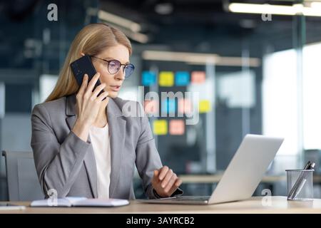 Eine fokussierte Geschäftsfrau in Brille, bei einem Anruf, arbeitet an ihrem Laptop an ihrem Schreibtisch. Das Büro hat Haftnotizen an der Wand. Stockfoto