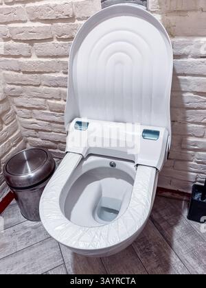 Die elegante, weiße Toilette hat einen minimalistischen Look mit einem einzigartigen Sitzdesign, das sich an einer Ziegelwand befindet Stockfoto