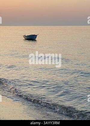 Kleines Holzboot, das auf dem ruhigen Adria-Wasser in der Nähe von Pineto herumtreibt und goldene Sonnenuntergangsreflexe über die sich kräuselnde Oberfläche wirft Stockfoto