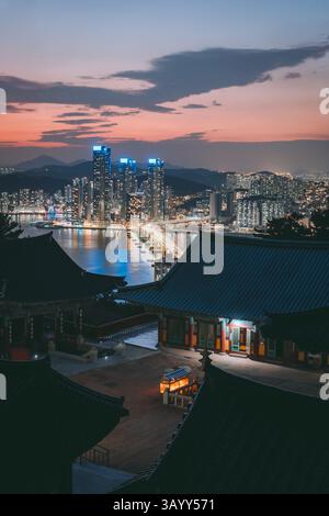 Abendliche Stadtlandschaft von Busan, Südkorea, mit traditionellem Tempel im Vordergrund und Wolkenkratzern, die entlang der Küste beleuchtet werden. Stockfoto