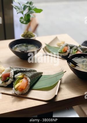 Traditionelle japanische Handbrötchen (Temaki-Sushi) gefüllt mit frischem rohem Fisch, Avocado und Gemüse, serviert auf einem Bambusblatt mit einer Seite Miso-Suppe. Stockfoto