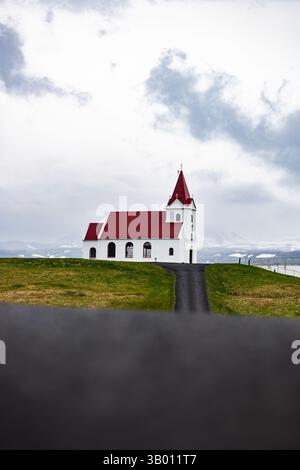 Isolierte weiße Kirche mit rotem Dach auf einem Hügel unter bewölktem Himmel, umgeben von isländischer Landschaft und weit entfernten schneebedeckten Gipfeln. Stockfoto