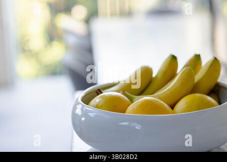 Die weiße Keramikschale hält Bananen und Orangen auf der Tischplatte in der modernen Küche nahe dem sonnendurchfluteten Fenster Stockfoto