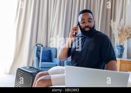 Afroamerikaner, der Smartphone hält und auf einem Laptop im Zimmer schreibt, mit Koffer Stockfoto