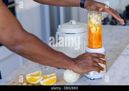 Afroamerikaner, der Frucht-Smoothie auf Marmorarbeitsfläche macht, mit Orangenscheiben Stockfoto