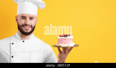 Glücklicher Konditor in Uniform, der Kuchen mit Beeren hält und eine gute Geste auf orangem Hintergrund zeigt Stockfoto