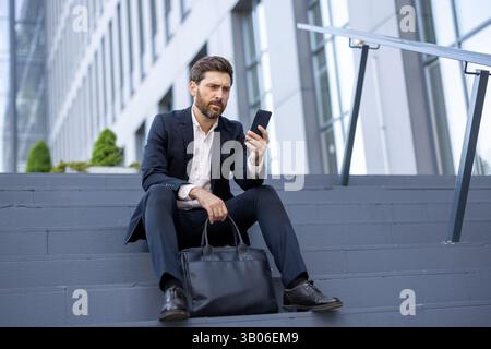 Ein besorgter Geschäftsmann im Anzug überprüft sein Handy, während er auf einer Treppe im Freien in der Nähe eines modernen Gebäudes sitzt. Er hält einen Aktenkoffer. Stockfoto