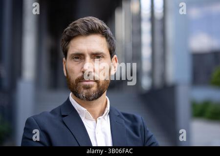 Ein gutaussehender, bärtiger Mann im Anzug posiert selbstbewusst und blickt mit einem komponierten Ausdruck direkt in die Kamera. Die urbane Kulisse deutet auf eine professionelle Umgebung hin. Stockfoto