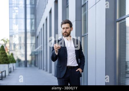 Ein Mann in einem Anzug steht vor einem Gebäude, in sein Handy eingedrungen. Er hält seine Hand in der Tasche und konzentriert sich auf den Bildschirm, während er seine Nachrichten überprüft. Stockfoto