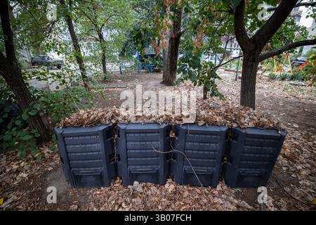 Auf dem Boden stehen in einer Reihe schwarze Kompostbehälter, die jeweils mit verschiedenen organischen Abfällen gefüllt sind: Blätter, Äste, Lebensmittelreste. Die Deckel sind geöffnet. Recycling Stockfoto