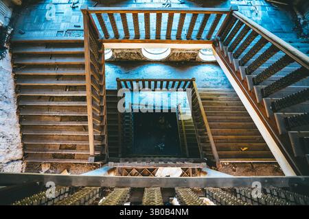 Blick von oben auf die Wendeltreppe in einem verlassenen Gebäude mit kunstvollem Geländer und runden Fenstern, geheimnisvolle Atmosphäre der verfallenen Struktur. Stockfoto