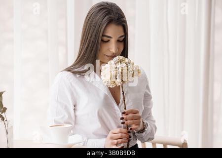 Junge kaukasische Frau riecht Hortensie, während sie drinnen mit Kaffeetasse am Tisch sitzt Stockfoto