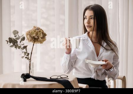 Elegante Frau mit Kaffee in der Nähe des Fensters und getrockneten Blumen in weißer Innenausstattung Stockfoto