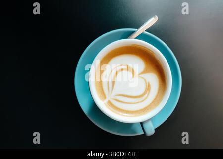 Tasse Cappuccino mit herzförmiger Latte Art auf Milchschaum in türkisfarbener Keramikschale mit Löffel auf dunklem Hintergrund. Draufsicht. Stockfoto