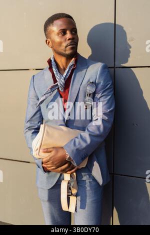 Eleganter Geschäftsmann mit Sonnenbrille in der Tasche, der eine Aktentasche hält und wegblickt, während er in der Nähe eines modernen Hotels steht Stockfoto