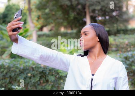 Die junge afroamerikanische Geschäftsfrau macht ein Selfie mit ihrem Smartphone im Park, macht ein Entengesicht und hält das Telefon mit der rechten Hand Stockfoto