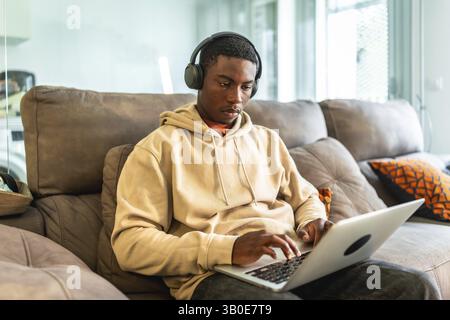 Junger schwarzer Mann, der Laptop und Kopfhörer benutzt, während er von zu Hause aus arbeitet oder studiert, auf einer bequemen Couch sitzt Stockfoto
