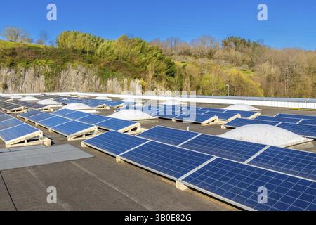 Reihen von Photovoltaik-Solarpaneelen, die Sonnenlicht auf einem Dach in Strom verwandeln, vor dem Hintergrund des blauen Himmels und eines bewaldeten Hügels Stockfoto